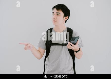 Selektive Fokusaufnahme eines jungen asiatischen Mannes, der ein lässiges T-Shirt in Salbeigrün, eine Smartwatch und einen Rucksack trägt, sucht mit dem Smartphone nach Wegbeschreibungen Stockfoto
