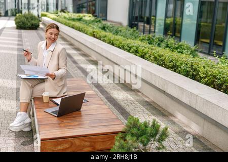 Analystin, die mit Dokumenten arbeitet und Laptop im Freien im modernen Gebäudehintergrund verwendet Stockfoto