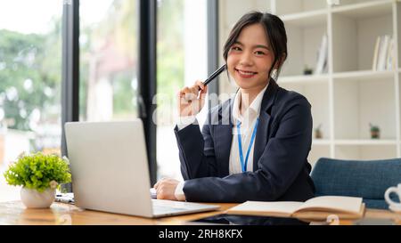 Eine attraktive und lächelnde asiatische Geschäftsfrau oder Buchhalterin in einem formellen Business-Anzug sitzt an ihrem Schreibtisch im Büro. Stockfoto