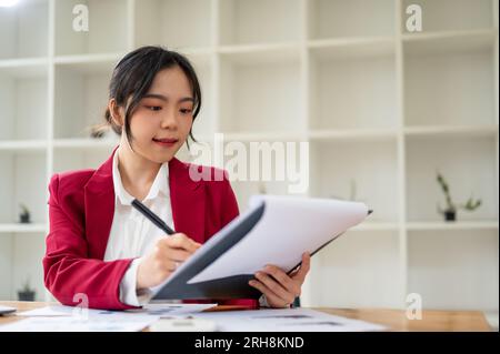 Eine schöne asiatische Geschäftsfrau in einem roten Business-Anzug konzentriert sich darauf, einen Geschäftsbericht an ihrem Schreibtisch im Büro zu prüfen. Stockfoto
