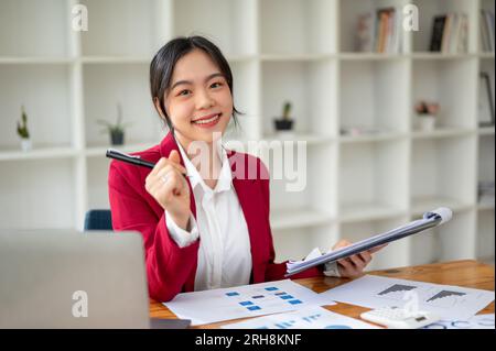 Ein Porträt einer fröhlichen und schönen asiatischen Geschäftsfrau in einem roten Business-Anzug sitzt an ihrem Schreibtisch mit Finanzberichten und einem Laptop und analysiert die Finanzen Stockfoto