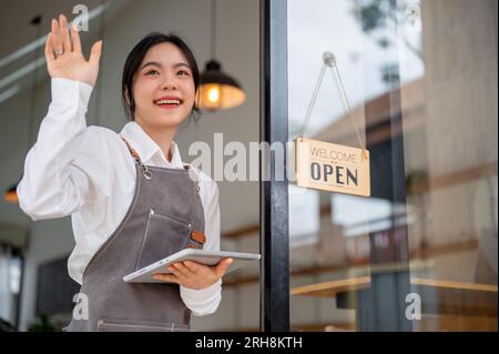 Eine hübsche und fröhliche asiatische Mitarbeiterin in einem Coffee Shop mit Schürze steht mit einem Tablet in der Hand vor der Eingangstür und verwaltet die Warteschlange für die Kunden Stockfoto