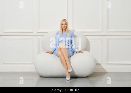 Hübsche Frau in hellblauem engen Strickkleid, die auf einem weichen Sessel sitzt und sich die Beine kreuzte Stockfoto