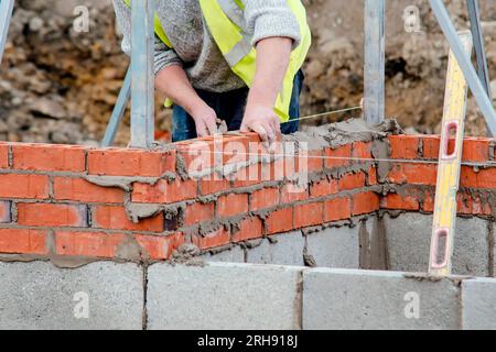 Hart arbeitender Maurer legt Ziegel auf Zementmischung auf der Baustelle. Bekämpfung der Wohnungskrise durch den Bau von erschwinglicheren Häusern Stockfoto