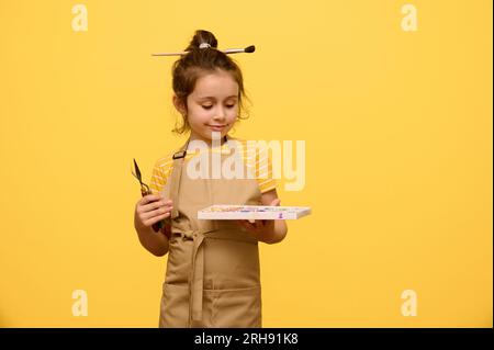 Porträt auf gelbem Hintergrund eines süßen kleinen Mädchens mit Malwerkzeugen, das Malerei lernt, in beigefarbener Schürze gekleidet und mit einem Pinsel im haarstyl Stockfoto