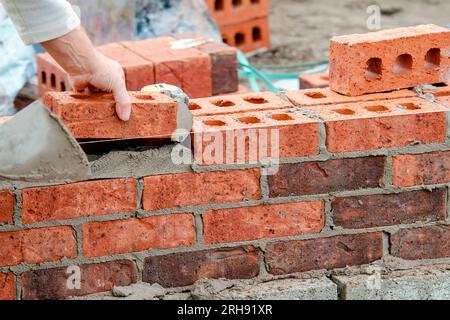 Hart arbeitender Maurer legt Ziegel auf Zementmischung auf der Baustelle. Bekämpfung der Wohnungskrise durch den Bau von erschwinglicheren Häusern Stockfoto