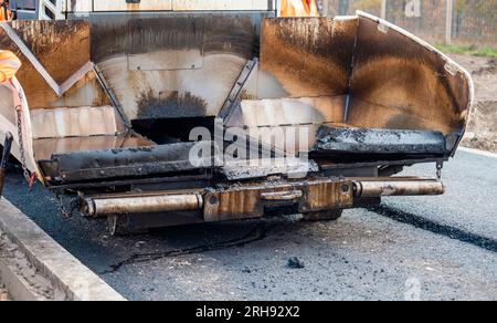 Asphaltfertiger mit heißem Asphalt, der neue Straßenoberflächen auf neuen Wohnbaustellen verlegt, und Straßenarbeiter in orangefarbenem Hi-viz als nächstes Stockfoto