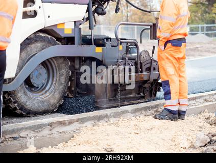 Asphaltfertiger mit heißem Asphalt, der neue Straßenoberflächen auf neuen Wohnbaustellen verlegt, und Straßenarbeiter in orangefarbenem Hi-viz als nächstes Stockfoto