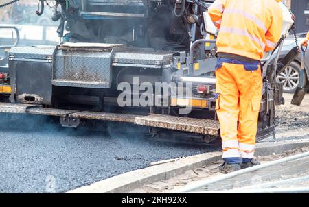 Asphaltfertiger mit heißem Asphalt, der neue Straßenoberflächen auf neuen Wohnbaustellen verlegt, und Straßenarbeiter in orangefarbenem Hi-viz als nächstes Stockfoto