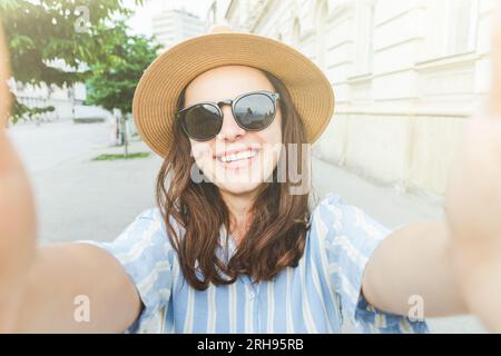 Junge lächelnde Frau mit Sonnenbrille und Hut, die auf der Straße steht und ein Selfie macht. Stockfoto