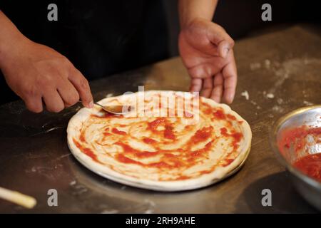Nahaufnahme eines Pizzamakers, der Tomatensauce auf Pizzateig verteilt Stockfoto