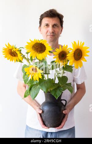 Ein Mann hält einen schwarzen Krug mit Sonnenblumen vor sich an eine weiße Wand Stockfoto