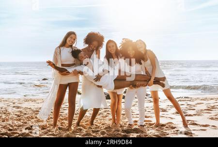 Fröhliche, vielfältige Frauen, die die Braut an den Händen halten, Frauen feiern die Hühnerparty am Strand an sonnigen Tagen, volle Länge Stockfoto