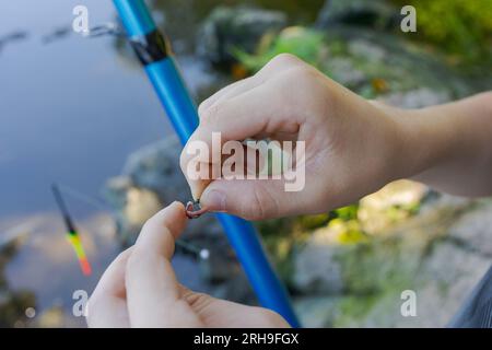 Nahaufnahme der Hände eines Teenagers, der Köder auf einen Angelhaken legt Sportfischen auf dem Fluss im Sommer. Stockfoto