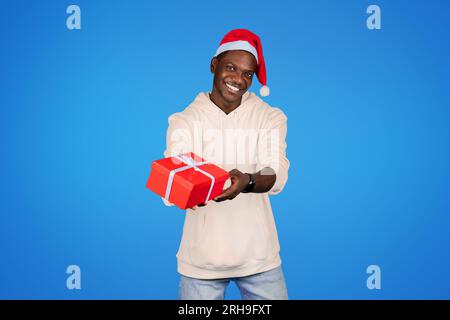 Lächelnder junger afroamerikanischer Mann im Sweatshirt, Weihnachtsmannmütze, Geschenkbox isoliert auf blauem Hintergrund, Studio Stockfoto