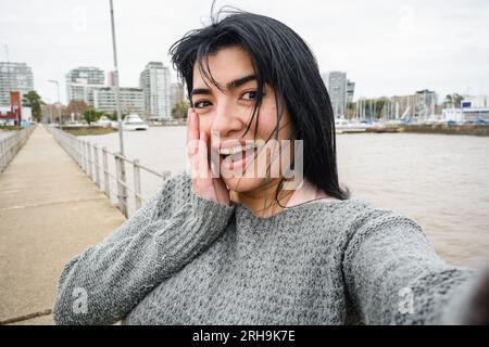 Die junge venezolanische lateinerin, die auf dem Pier über dem Fluss in buenos aires steht, lacht und macht lustiges Selfie-Portrait mit ihrem Telefon, ihrem Technologiekonzept, Stockfoto