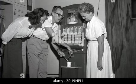 1950er, historisch, in einem Umkleidebereich, beobachten zwei Damen, wie ein männlicher Kollege an einem Automaten, der „Snack Bar; USA“, ein Getränk bekommt. Getränke verfügbar, schwarzer Kaffee, Kaffee mit Sahne, heiße Coca & Chicken Bouillon Suppe. Jede Packung heißes Getränk kostet 10 Cent und man kann ein oder zwei Cent einstecken! Einer fügte dann heißes Wasser hinzu. Die erste echte kommerzielle Nutzung von Verkaufsautomaten war Ende der 1880er Jahre, um Kaugummi auf Bahnsteigen zu verkaufen, und in den frühen 1900er Jahren wurden alkoholfreie Getränke angeboten. Kaffee- und Heißgetränk-Automaten erschienen seit den späten 1940er Jahren. Stockfoto