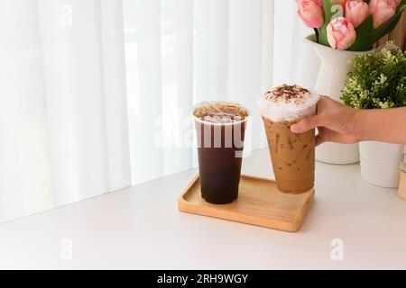 Ice americano und Ice Cappuccino Kaffee und ein Holztablett auf einem weißen Tisch im Innenbereich mit weißem Vorhang mit Blumen dekoriert. Stockfoto