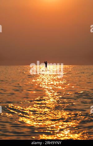 Farbenfroher Himmel bei Sonnenuntergang mit der Silhouette eines Mannes, der auf dem Paddleboard auf dem Meer surft. Stockfoto