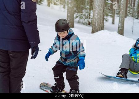 Ein kleiner Junge lernt, auf einer Skipiste auf dem Snowboard zu fahren. Winterurlaub. Kiefernwald im Hintergrund. Vater unterrichtet Sohn Snowboarden Stockfoto