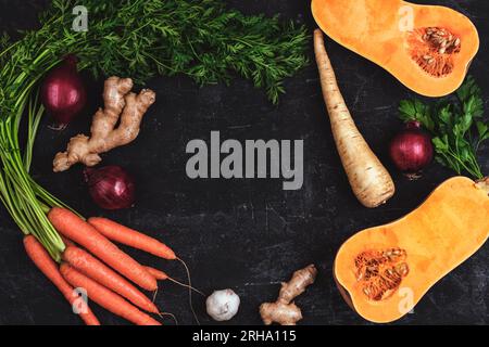 Rahmen aus Gemüse, Kürbis, Karotten, Ingwer, Zwiebeln, Pastinake und Petersilie auf schwarzem strukturiertem Hintergrund. Gesundes rohes veganes Essen. Draufsicht, flach, Co Stockfoto