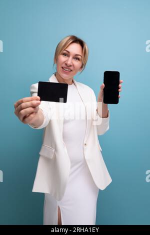 Eine selbstbewusste junge europäische Frau in weißer Jacke und weißem Kleid wirbt mit einer Bankkarte und einem Smartphone Stockfoto