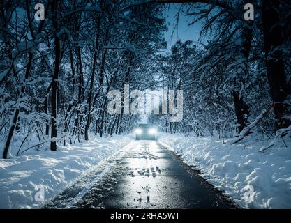 Bei Nacht vor einem Auto mit hellen Scheinwerfern stehen. Symmetrischer Schuss in kalten Tönen der Vorderseite eines Autos auf schmalem, schneebedecktem Weg durch den Wald Stockfoto