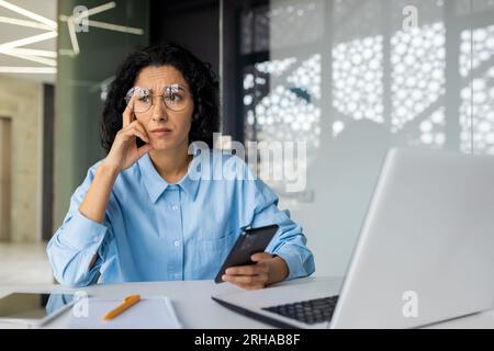 Traurig verärgerte Geschäftsfrau am Arbeitsplatz, hispanische Frau enttäuscht von Arbeitsergebnissen im Büro, weibliche Mitarbeiterin mit Laptop, Smartphone in der Hand. Stockfoto