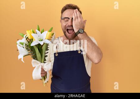 Ein Mann mittleren Alters mit Bartflorist, der Blumen hält, die ein Auge mit der Hand bedecken, selbstbewusstes Lächeln im Gesicht und überraschende Emotionen. Stockfoto