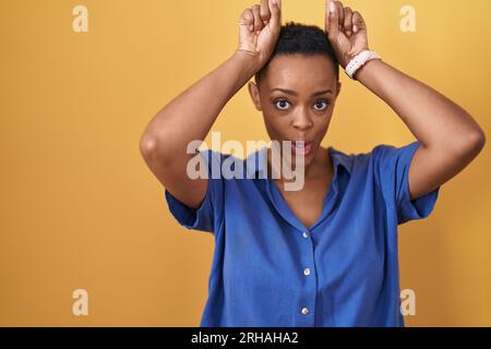 Eine afroamerikanische Frau, die auf gelbem Hintergrund steht und eine lustige Geste macht, mit dem Finger über dem Kopf wie Bullenhörner Stockfoto