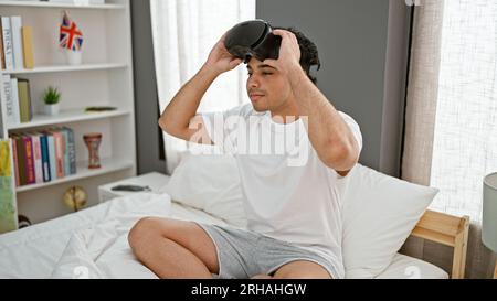 Ein junger latino, der im Bett sitzt und eine Virtual-Reality-Brille im Schlafzimmer trägt Stockfoto