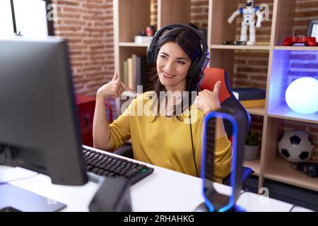 hispanische Frau im mittleren Alter, die Videospiele spielt und Kopfhörer verwendet, die selbstbewusst aussehen und mit einem Lächeln im Gesicht auf sich zeigen, stolz und glücklich mit den Fingern. Stockfoto