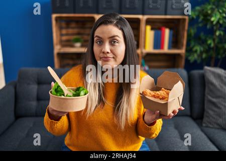 Junge hispanische Frau mit gesundem Salat und gebratenen Hähnchenflügeln, die lächelnd zur Seite blickt und wegstarrt und denkt. Stockfoto