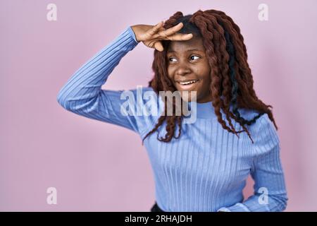 Afrikanische Frau, die über rosa Hintergrund steht, sehr glücklich und lächelnd, mit der Hand über dem Kopf weit weg. Suchbegriff. Stockfoto
