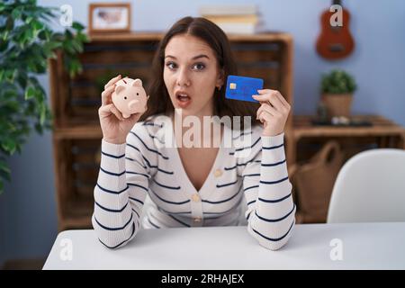 Junge hispanoamerikanerin mit Sparschwein und Kreditkarte, ahnungslos und verwirrter Ausdruck. Zweifelhaftes Konzept. Stockfoto