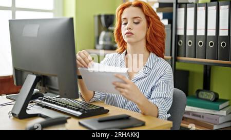 Junge rothaarige Geschäftsangestellte, die im Büro einen Briefumschlag zum Öffnen des Computers benutzt Stockfoto