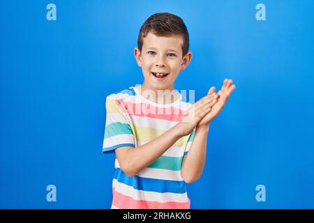 Junger Kaukasier, der über blauem Hintergrund steht, klatscht und applaudiert fröhlich und fröhlich, lächelt stolze Hände zusammen Stockfoto