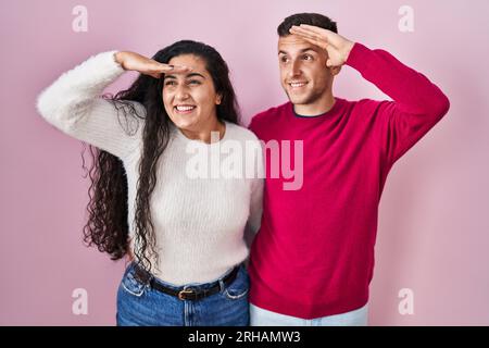 Das junge hispanische Paar, das über einem rosa Hintergrund stand, war sehr glücklich und lächelte und schaute mit der Hand über dem Kopf weit weg. Suchkonzept. Stockfoto