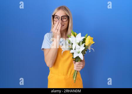 Junge weiße Frau, die Blumenschürze trägt, Blumen hält, lacht und kichert peinlich, verdeckt den Mund mit Händen, Klatsch und Skandal Stockfoto