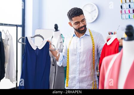 Junger arabischer Mann, Schneider, lächelnd, hält selbstbewusst T-Shirts in der Schneiderei Stockfoto