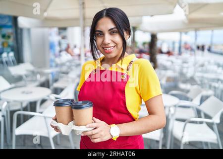 Junge schöne arabische Kellnerin lächelt selbstbewusst und hält Kaffee zum Mitnehmen auf der Terrasse des Cafés Stockfoto