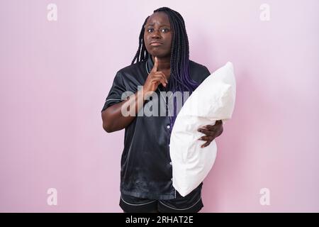 Junge afrikanische Frau, die Pijama-Kissen trägt und denkt über Zweifel mit dem Finger am Kinn nach und schaut hoch und wundert sich Stockfoto