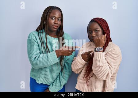 Zwei afrikanische Frauen, die auf blauem Hintergrund stehen, in Eile auf die Zeit zeigen, ungeduldig, mit entspanntem Ausdruck in die Kamera schauen Stockfoto