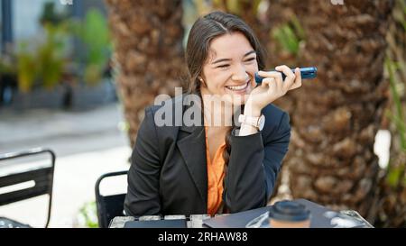 Eine junge, wunderschöne hispanische Geschäftsfrau, die eine Sprachnachricht per Smartphone sendet und auf der Terrasse des Cafés lächelt Stockfoto