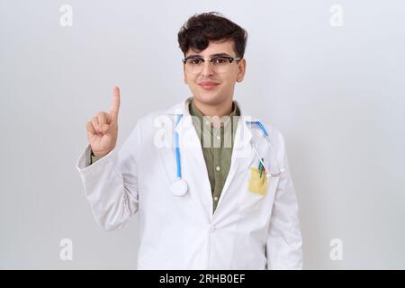 Junger, nicht binärer Mann in Arztuniform und Stethoskop, der mit Finger Nummer eins nach oben zeigt und dabei selbstbewusst und glücklich lächelt. Stockfoto