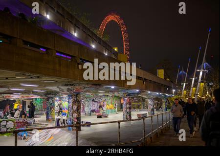 LONDON - 22. April 2023: Touristen schlendern vorbei an einem Graffiti-überdachten Skatepark auf der Southbank, London, während die orangefarbene Beleuchtung des London Eye ein ca. Stockfoto