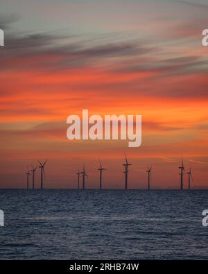 Weit entfernter Windpark bei Sonnenaufgang. Die Turbinen liegen in einer ruhigen Nordsee und der Himmel ist farbenfroh orange und blau und teilweise bewölkt. Stockfoto