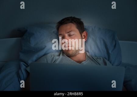 Junger Mann, der vor einem Laptop in einem Bett zu Hause schläft, müde nach dem Arbeitstag. Der Mann schläft ein, während er nachts auf seinem Computer Filme ansieht Stockfoto