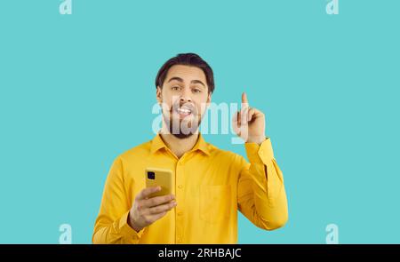 Bärtiger kaukasischer Mann mit Handy zeigt den Finger nach oben, wenn er eine tolle Idee hat, steht im Studio Stockfoto