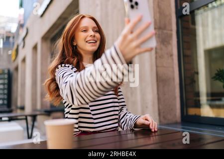 Nahaufnahmen von unten, bei denen eine glückliche junge Frau mit einem Lächeln auf den Smartphone-Bildschirm schaut, Videoanrufe im Freien genießt und im urbanen Café sitzt. Stockfoto